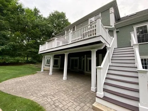 A two-story house with a green exterior features an elevated deck with white railings, stairs leading down, and a paved patio area underneath. Trees and grass are visible in the background. porch construction