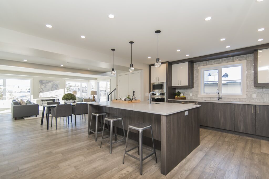 Modern kitchen with wood flooring, dark cabinets, large island with three stools, pendant lights, and open view to a dining area and living room with large windows—perfectly crafted by a trusted kitchen remodel contractor service.